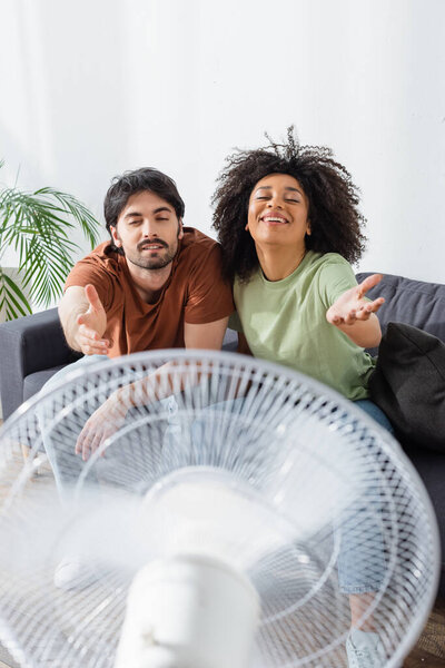 happy interracial couple sitting on couch with outstretched hands near blurred electric fan in living room 