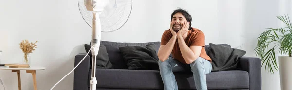 Homem Barbudo Feliz Sentado Sofá Perto Ventilador Elétrico Borrado Banner — Fotografia de Stock