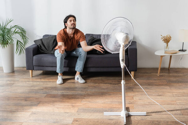 pleased and bearded man sitting on couch near blurred electric fan 