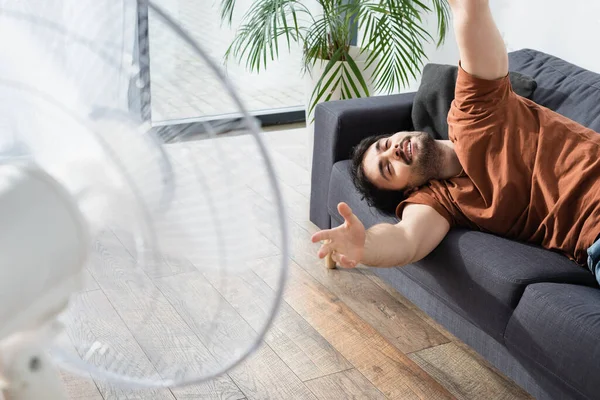 Tevreden Bebaarde Man Liggend Bank Buurt Wazig Elektrische Ventilator — Stockfoto