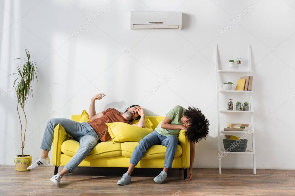 interracial couple suffering from heat on yellow couch in modern living room 