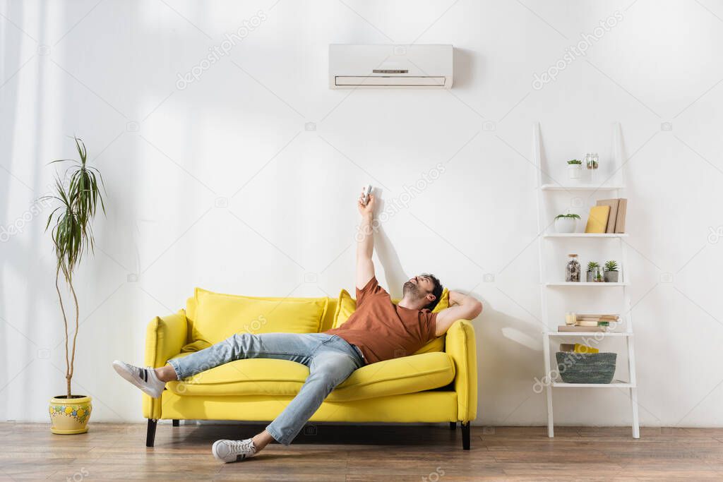 man using remote controller near air conditioner while suffering from heat in living room