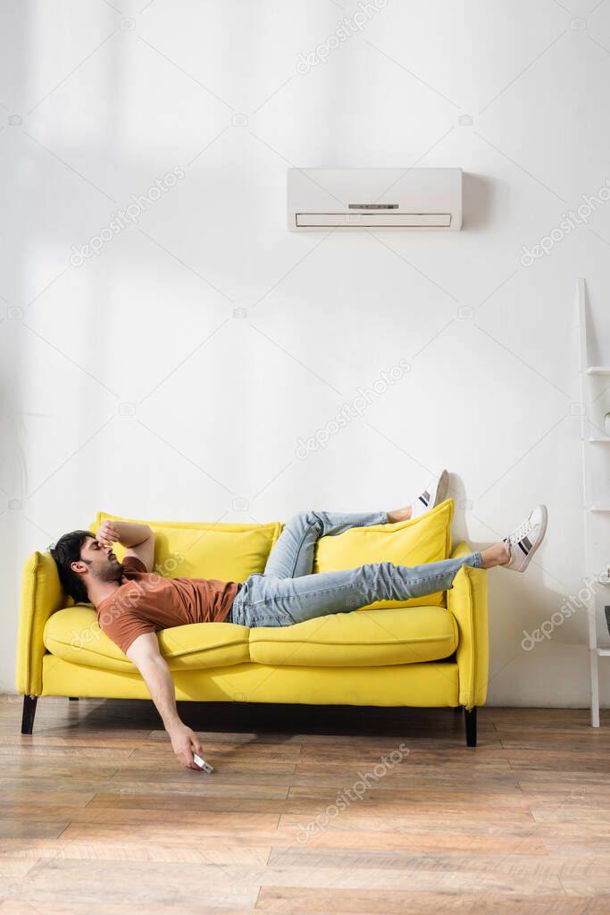 man holding remote controller while lying on yellow couch and suffering from heat in living room