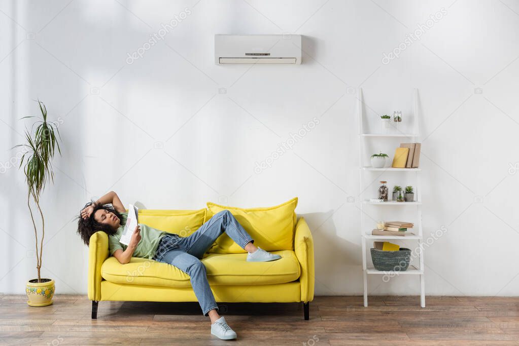 african american woman waving with newspaper while lying on yellow couch and suffering from heat