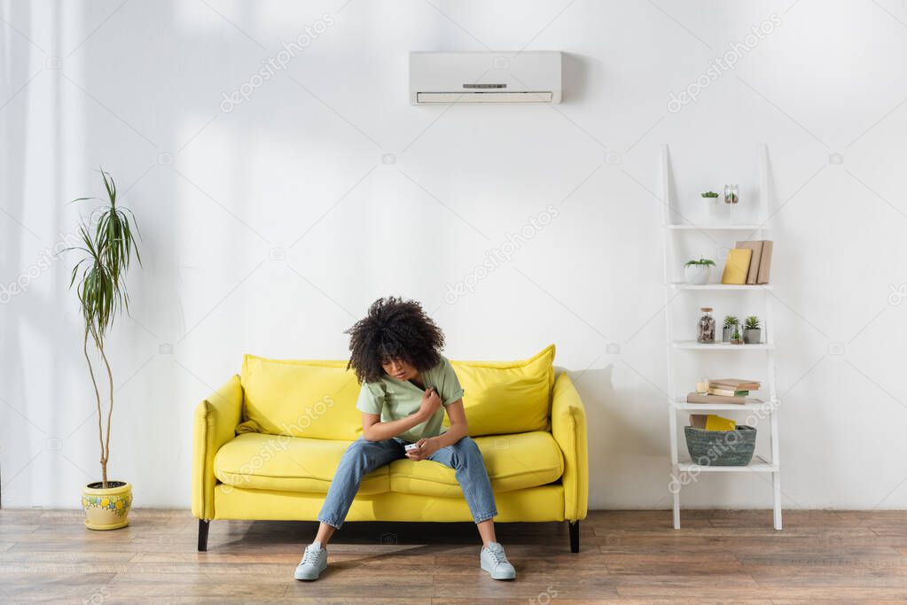 displeased african american woman holding remote controller while sitting on yellow sofa and suffering from heat