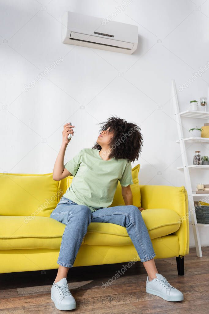 Disappointed african american woman holding remote controller while sitting on yellow couch and suffering from heat