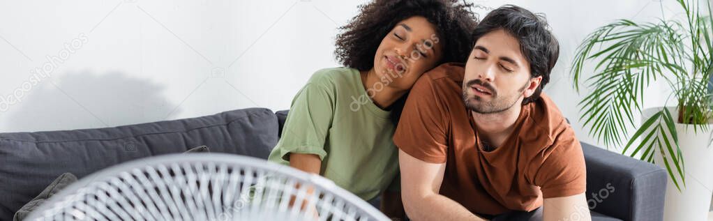 pleased interracial couple sitting on couch near blurred electric fan in living room, banner 