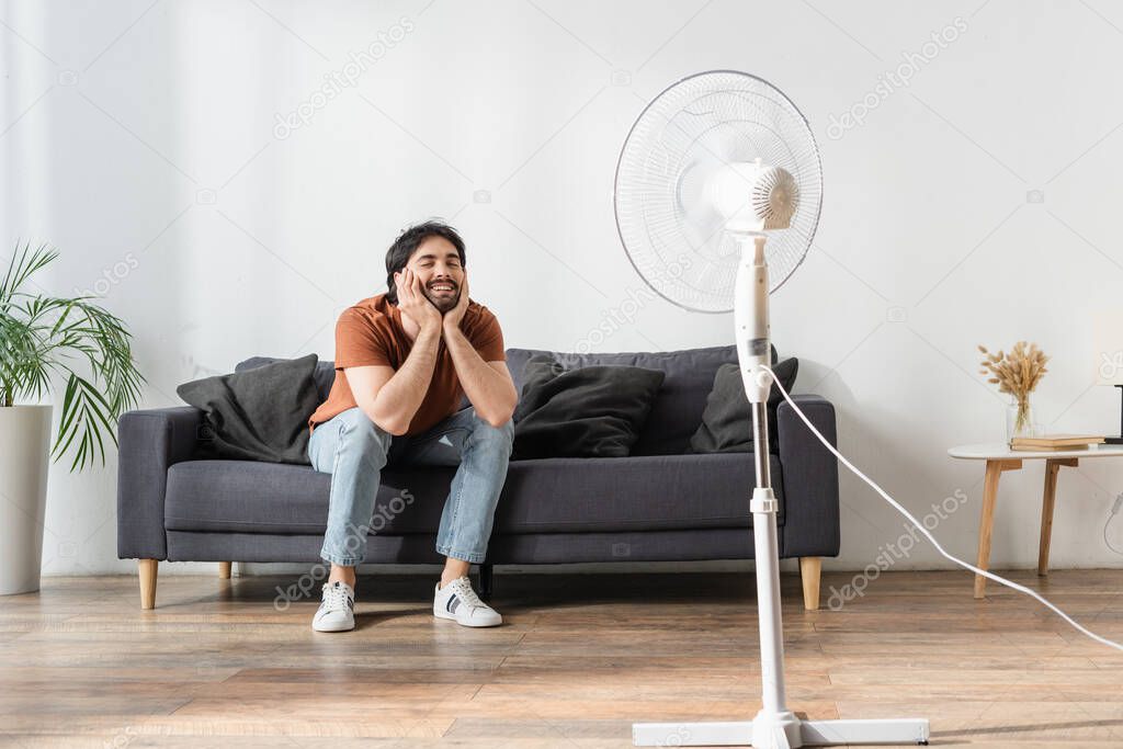 happy bearded man sitting on couch near blurred electric fan 