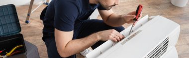 cropped view of bearded handyman holding screwdriver while fixing broken air conditioner, banner clipart
