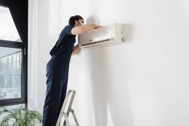 repairman in medical mask standing on ladder and fixing air conditioner clipart