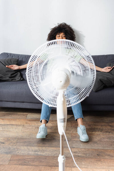 happy african american woman resting on couch near blurred electric fan