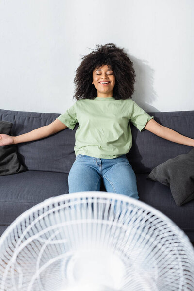 cheerful african american woman resting on couch near blurred electric fan