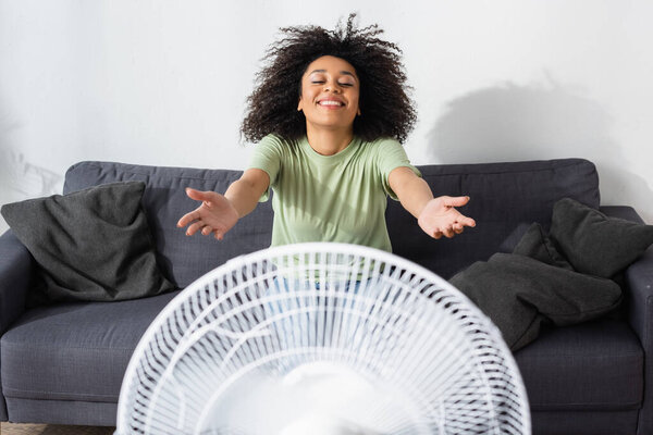 joyful african american woman sitting with outstretched hands on couch near blurred electric fan