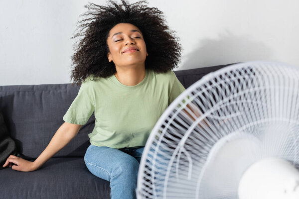 joyful african american woman sitting on couch near blurred electric fan