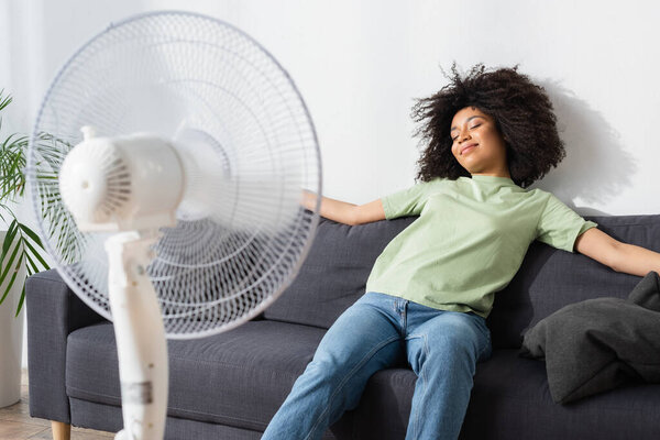 pleased african american woman resting on couch near blurred electric fan