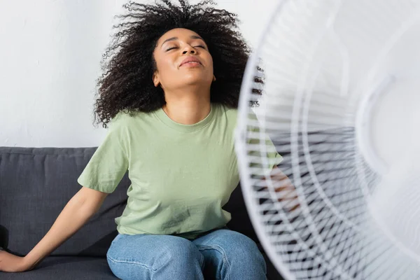 Kudrnatý Africký Americký Žena Sedí Gauči Blízkosti Rozmazané Elektrický Ventilátor — Stock fotografie