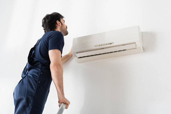 handyman in overalls standing on ladder while fixing broken air conditioner 