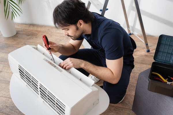 bearded handyman holding screwdriver while fixing broken air conditioner in living room