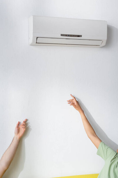 cropped view of multiethnic couple reaching air conditioner while suffering from heat in summer