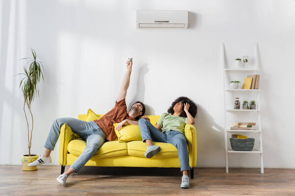 man holding remote controller near modern air conditioner and african american girlfriend suffering from heat