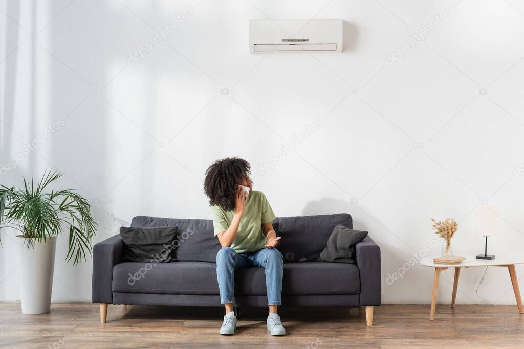 displeased african american woman talking on smartphone and looking at air conditioner in living room 