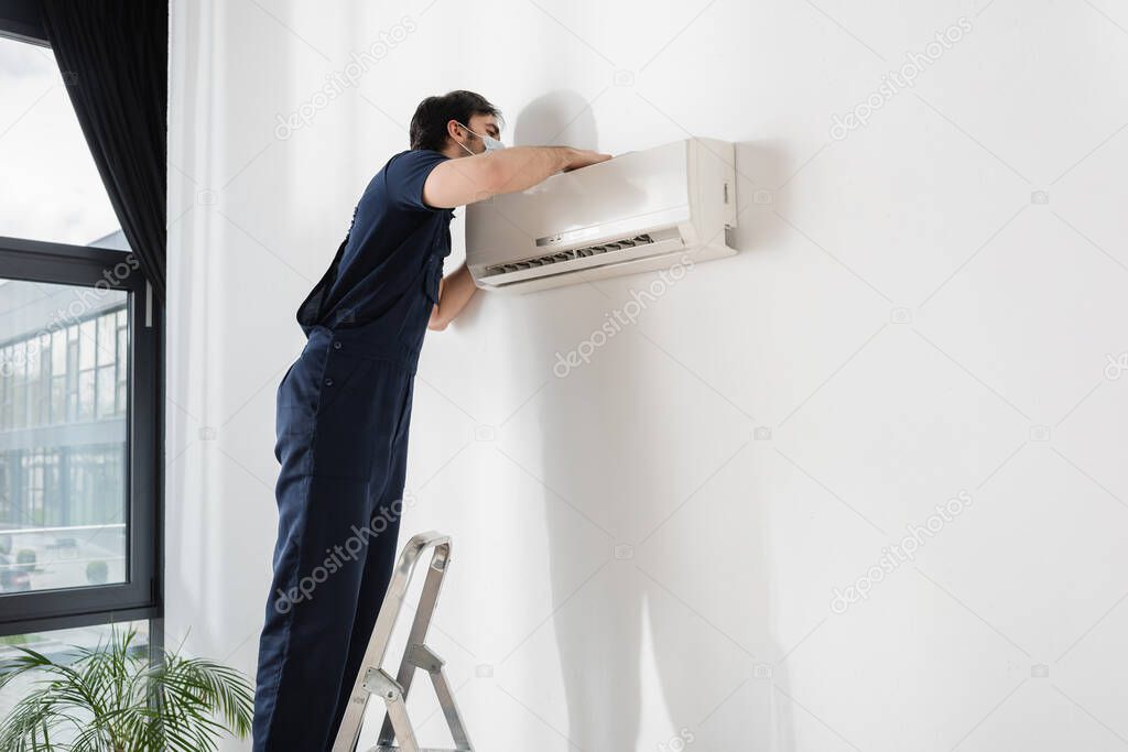 repairman in medical mask standing on ladder and fixing air conditioner