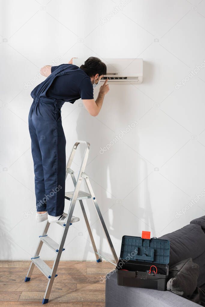 repairman in medical mask standing on metallic ladder and fixing air conditioner