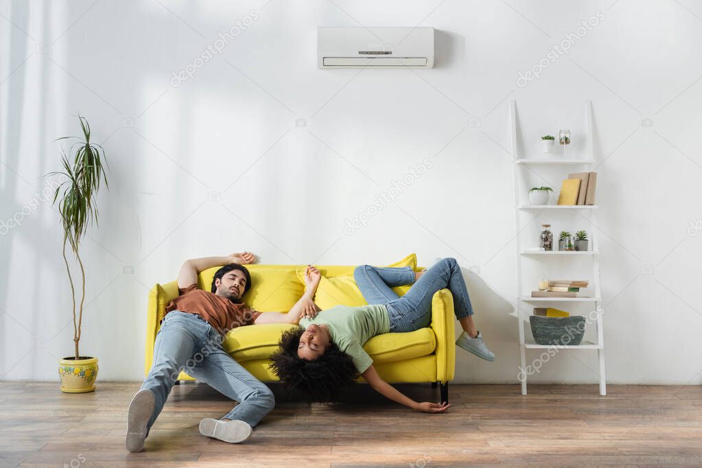 exhausted young multiethnic couple lying on yellow sofa and suffering from heat in summer