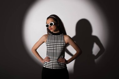 lighting on stylish model in sunglasses posing with hands on hips on grey  clipart