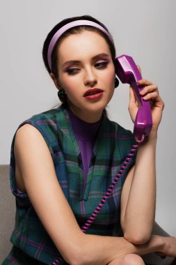 Gri koltukta izole bir şekilde otururken düşünceli genç bir kadın retro telefonda konuşuyor.