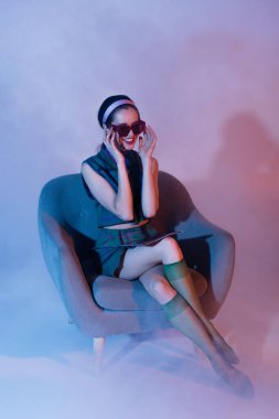 Neşeli genç bir kadın güneş gözlüğünü ayarlıyor ve menekşe dergisi ile koltukta oturuyor.