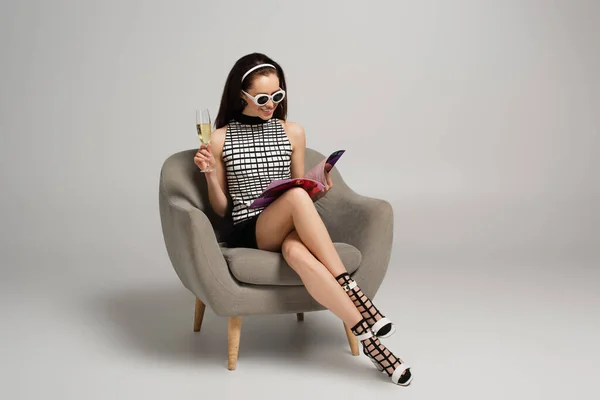 坐在灰色扶手椅上 戴着复古太阳镜 一边看杂志 一边举着香槟的年轻女子微笑着 — 图库照片