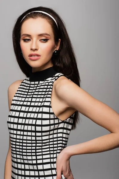 young model in headband, earrings and retro black and white outfit posing with hand on hip isolated on grey