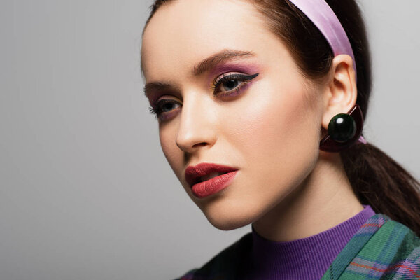 молодая женщина в фиолетовом повязке и серьге глядя в сторону изолированы на серый 