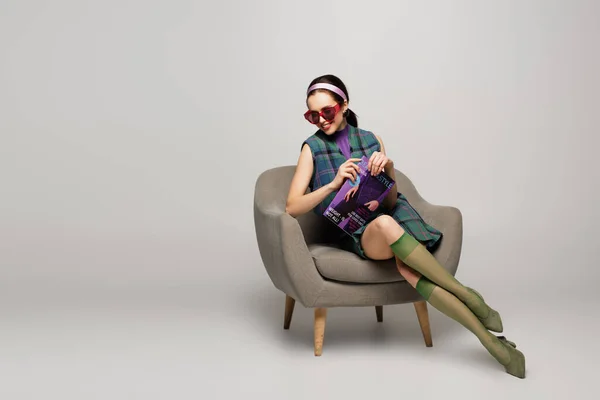 Zufriedene Junge Frau Mit Sonnenbrille Und Magazin Sessel Auf Grau — Stockfoto