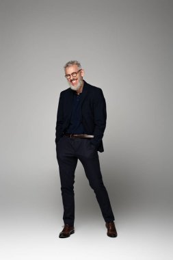 Orta yaşlı, gözlüklü, takım elbiseli, gülümseyen ve elleri gri cepli bir adam.