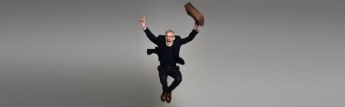 full length of amazed man with grey hair holding leather bag while levitating on grey, banner clipart