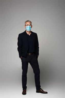 Gri saçlı, gözlüklü ve tıbbi maskeli, elleri gri cepli olgun bir adam. 