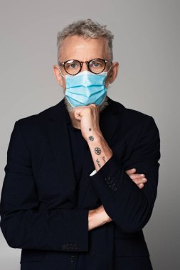 Dövmeli ve olgun, gri saçlı, gözlüklü ve maske takan gri saçlı bir adam. 