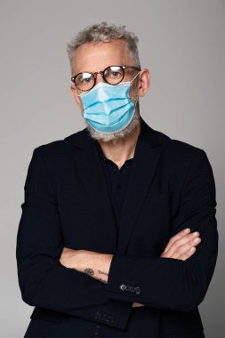 Gri saçlı, gözlüklü ve tıbbi maskeli olgun bir adam gri üzerine izole edilmiş poz veriyor. 