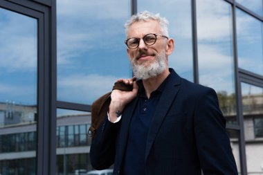 Gülümseyen, gri saçlı, deri çantalı olgun bir adam binanın yanında dikilirken. 