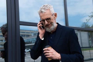 Kafası karışık bir iş adamı elinde kağıt bardakla modern binanın yakınında akıllı telefondan konuşuyor. 
