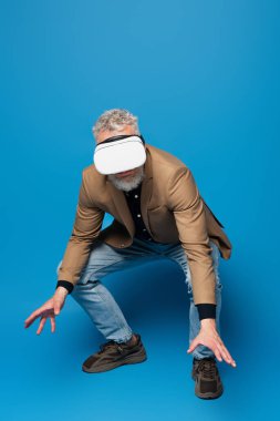 full length of middle aged man with grey hair in vr headset squatting and gesturing on blue  clipart