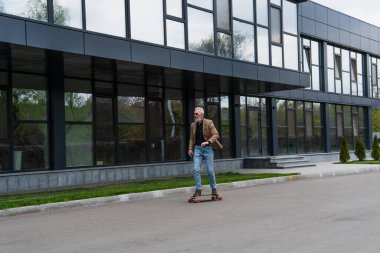 Güneş gözlüklü ve kot pantolonlu orta yaşlı bir adam dışarıda uzun tahta sürüyor. 