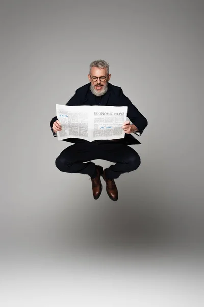full length of middle aged man in glasses and suit reading economic news while levitating on grey