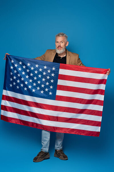 full length of bearded middle aged man standing with flag of america on blue 