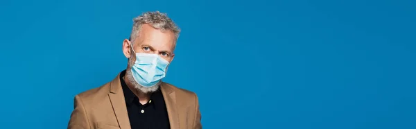 middle aged man in protective medical mask looking at camera isolated on blue, banner
