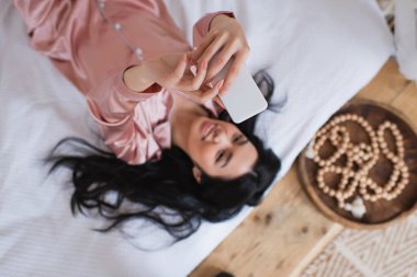 Yatakta uzanmış cep telefonuyla selfie çeken ipek pijamalı, gülümseyen Asyalı bir kadın.