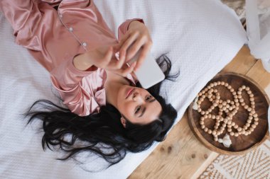 İpek pijamalı, yatakta uzanmış ve cep telefonuyla selfie çeken Asyalı genç bir kadın.