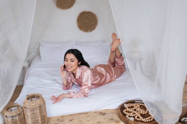 Pembe ipek pijamalı genç Asyalı kadın yatakta uzanıyor ve yatak odasında cep telefonuyla konuşuyor.